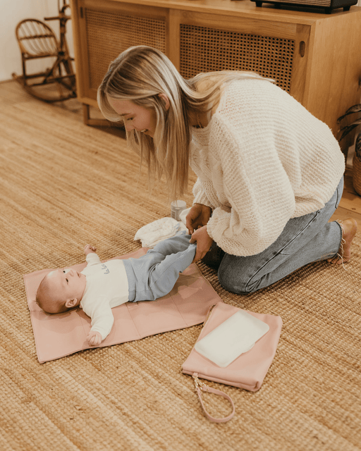 A baby is being changed on the floor, laying comfortably on a pretty pink change mat.
